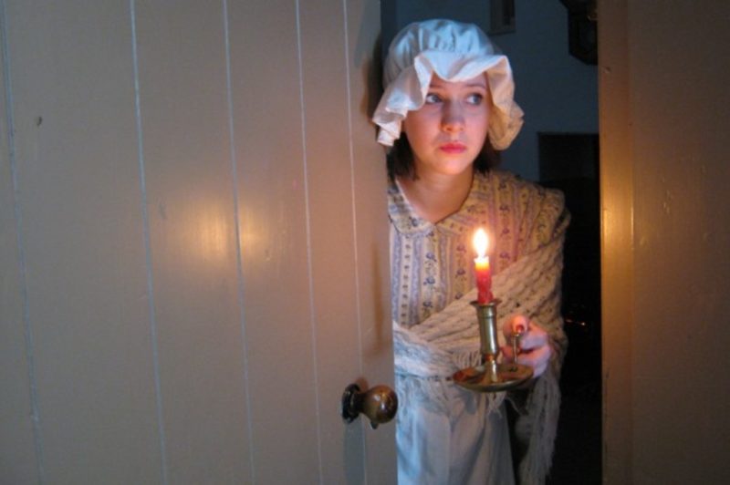 Scared girl in Victorian clothes peering round an open door with a candle.